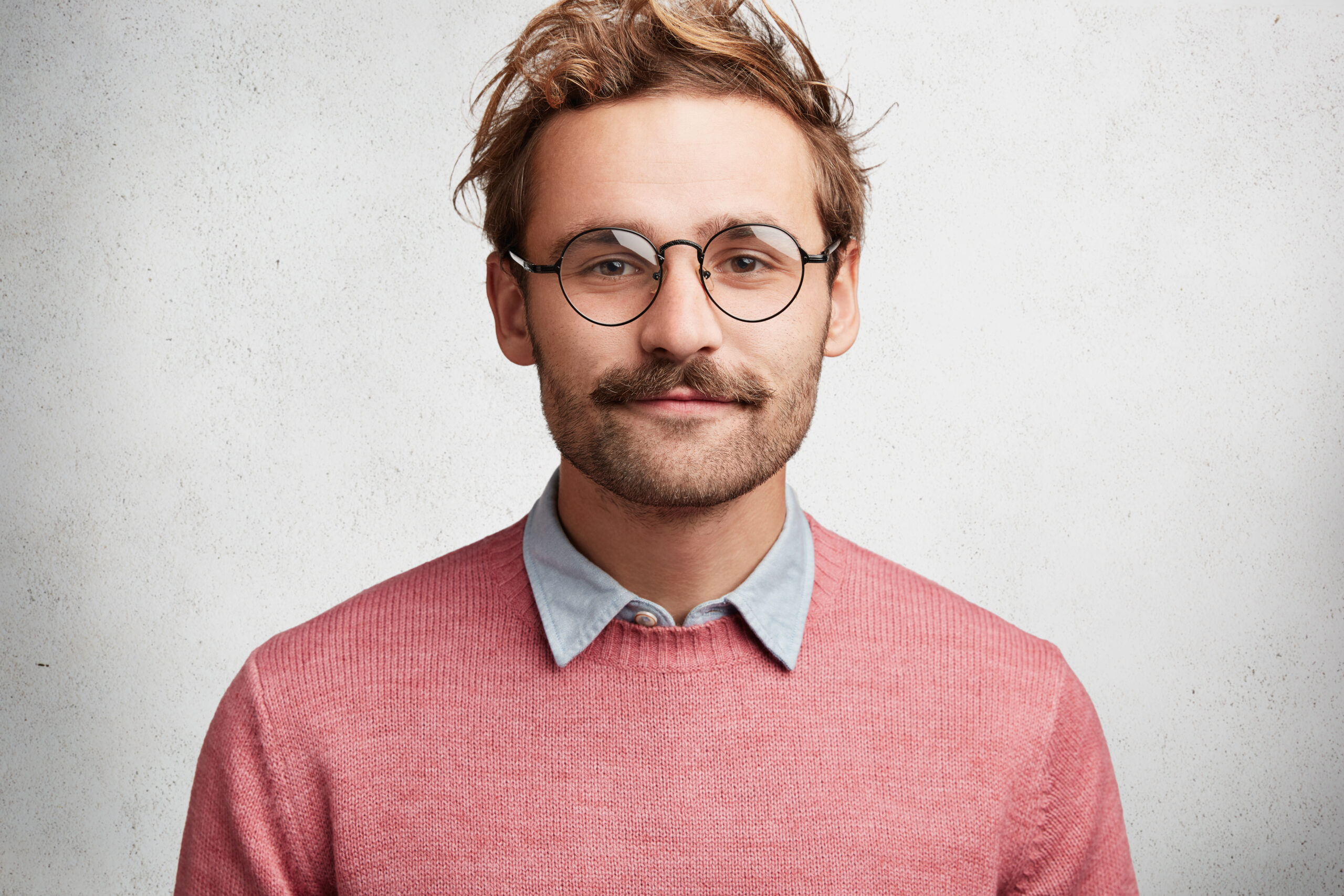 Indoor shot of delightful bearded young male professor wears spectalces, attends seminar, listens to lecture attentively, dressed in shirt and sweater, isolated over white concrete background